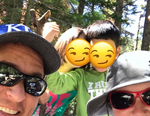 At the left bottom of the picture, a man's smiling face is partially visible. He has brown hair and is wearing a KU baseball cap and black sunglasses. At the lower right of the picture, a woman's face is visible. She is wearing a tan hiking hat and pink sunglasses with black lenses. Behind them, two children stand facing the camera with their heads together. The child on the left has brown hair and brown skin, and is wearing a teal t-shirt with the word "BE" in hot pink print. The child on the right has black hair and brown skin, and is wearing a long-sleeved green t-shirt with white print on it (the words can't be made out). The kids' faces are covered with half-smile emojis to protect their identities. Behind them all is a forest of tall pine trees.